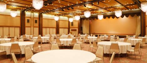 A banquet hall with round tables and chairs, a stage, and chandeliers.