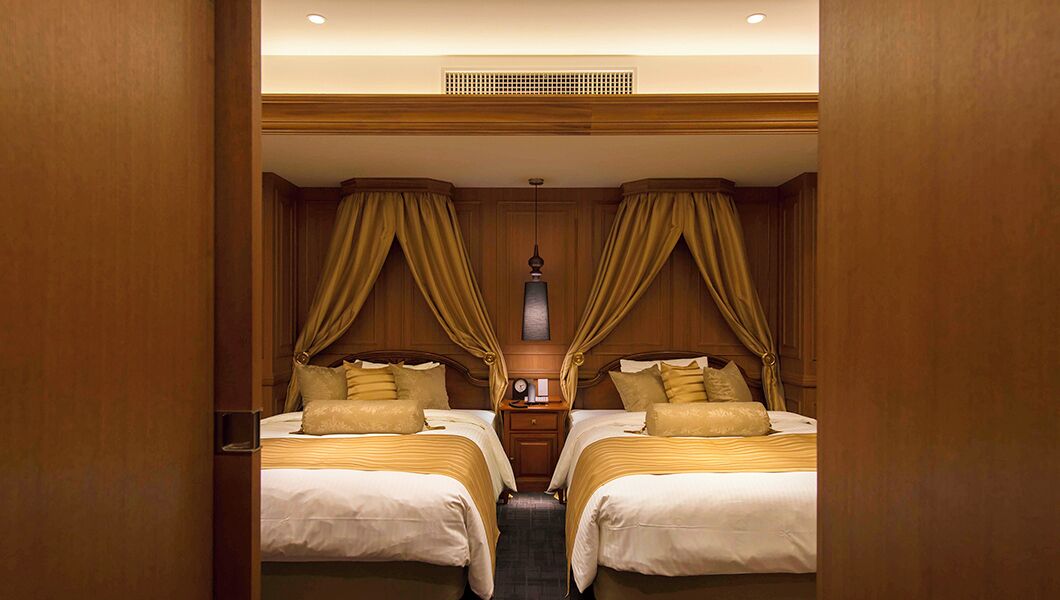 Quarto de hotel com duas camas, painéis de madeira, uma ventilação no teto e um lustre.