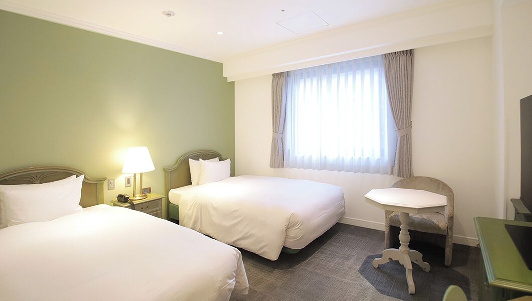 A hotel room with two beds, a desk, a chair, a lamp, and a window with curtains.