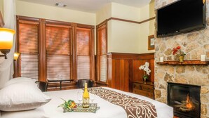A hotel room with a large bed, wooden blinds, a stone fireplace, a mounted TV, and a dining area with a table and chairs. - Hotel Napa Valley, an Ascend Collection Hotel (Napa)