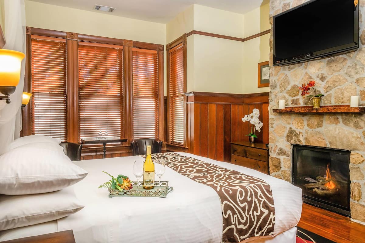 A hotel room with a large bed, wooden blinds, a stone fireplace, a mounted TV, and a dining area with a table and chairs.