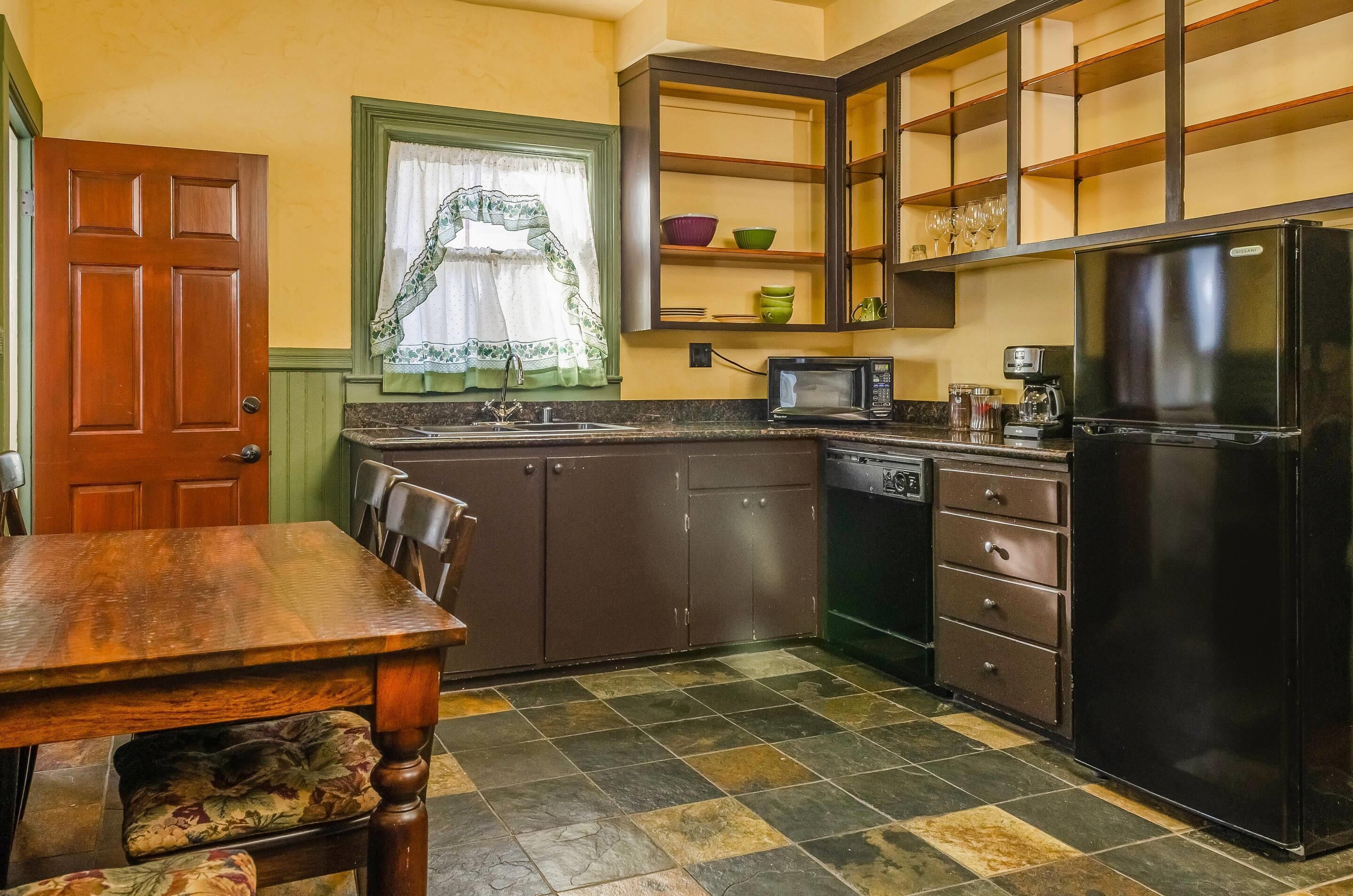 A kitchen with a wooden table, cabinets, a refrigerator, a microwave, and a window with a curtain.