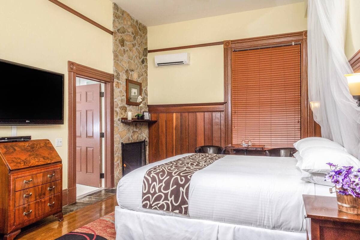 A hotel room with a large bed, a flat-screen TV, a wooden dresser, a stone accent wall, and a fireplace.