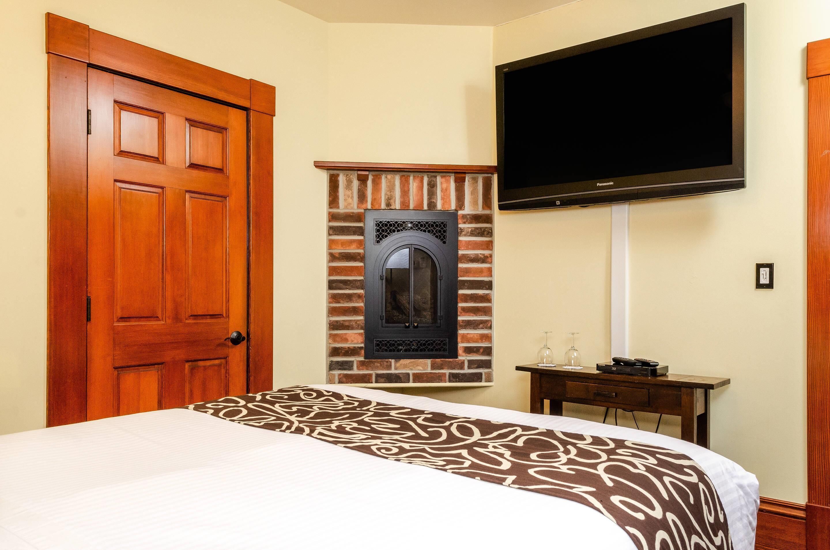 A hotel room with a bed, a wooden door, a brick fireplace, a television, and a small table with a glass.