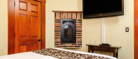 A hotel room with a bed, a wooden door, a brick fireplace, a television, and a small table with a glass.