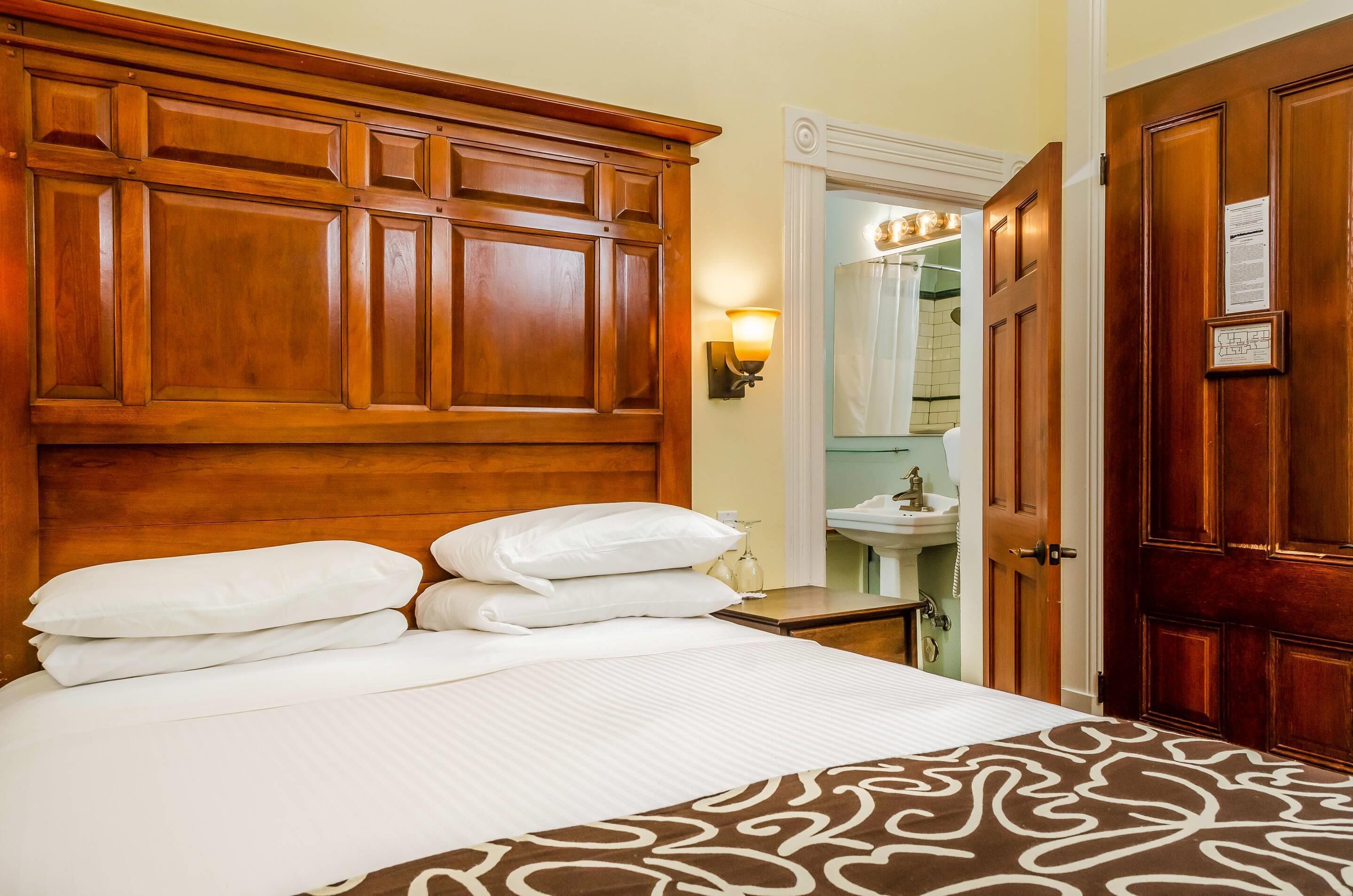 A hotel room with a large wooden headboard, a bedside table with a lamp, and a bathroom visible through an open door.