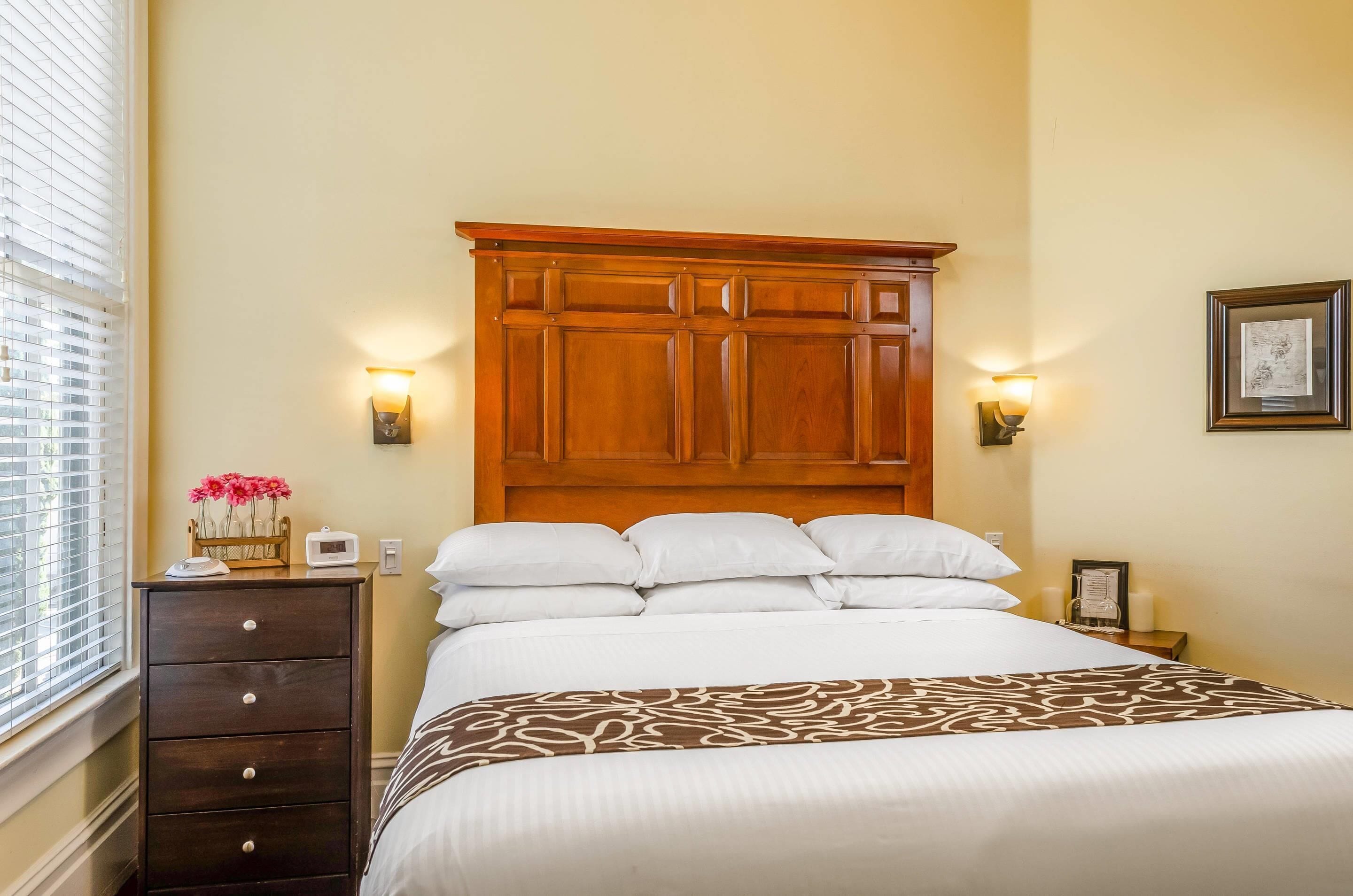 A neatly made bed with a patterned throw, a wooden headboard, bedside lamps, a nightstand with a framed picture, and a dresser with drawers.