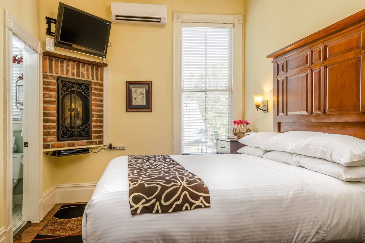 A hotel room with a brick fireplace, a flat-screen TV mounted above it, a large bed with white bedding, a wooden headboard, and a window with blinds.