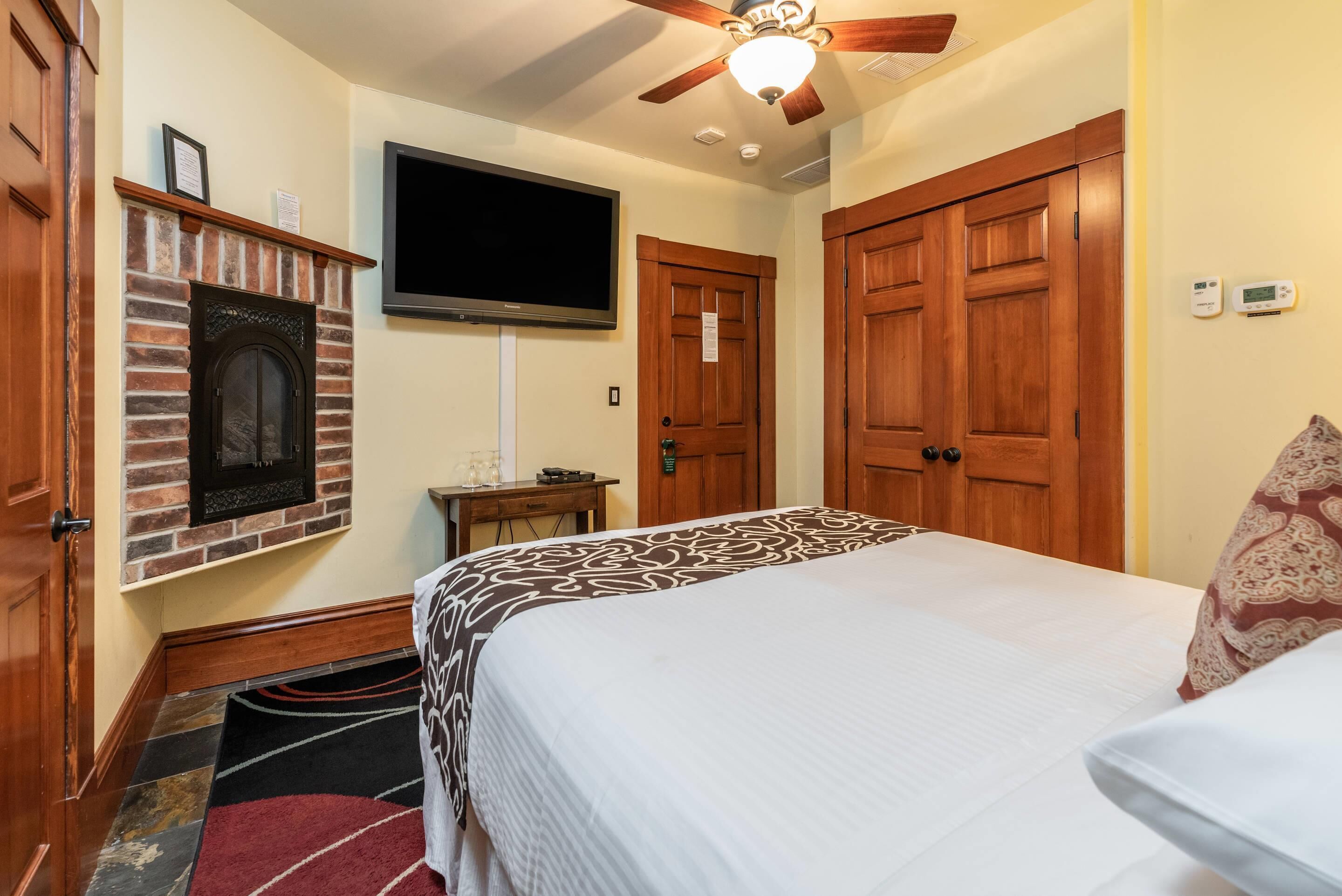 A hotel room with a brick fireplace, a flat-screen TV, wooden doors, and a bed with a patterned bedspread.