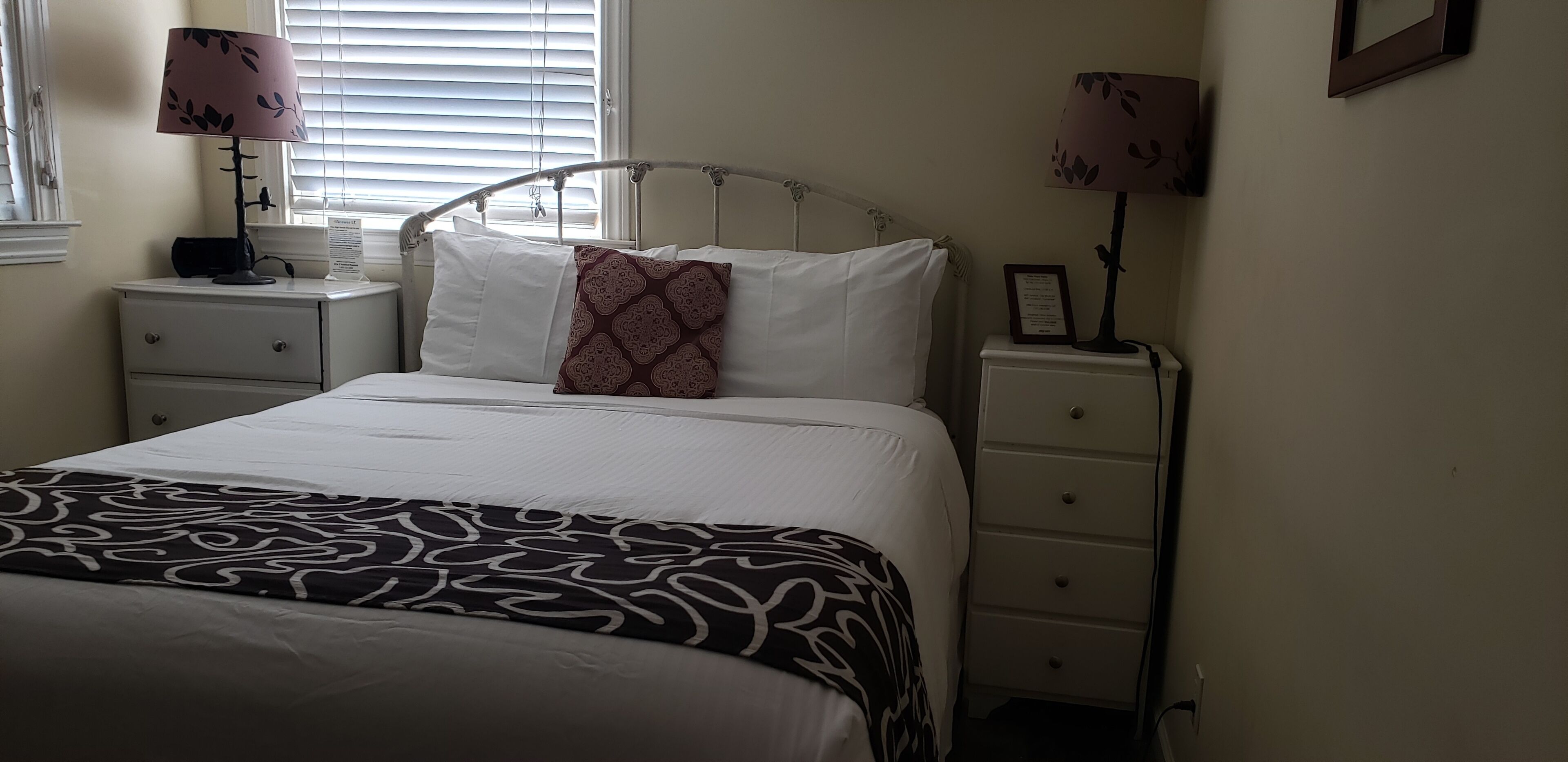Cama bem arrumada com colcha estampada, duas luminárias de cabeceira, mesa de cabeceira e um quadro na parede.