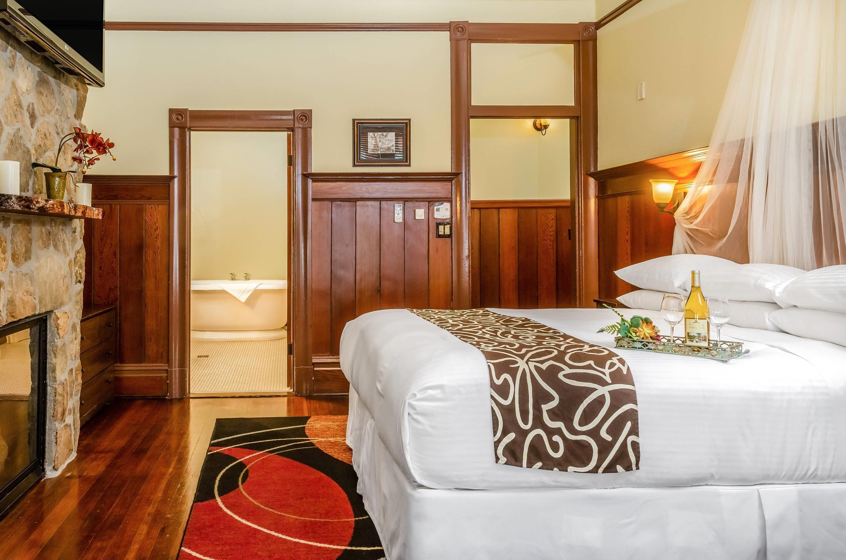 Una habitación de hotel con cama, mesita de noche, lámpara, ventana con cortina y puerta que da a un baño.