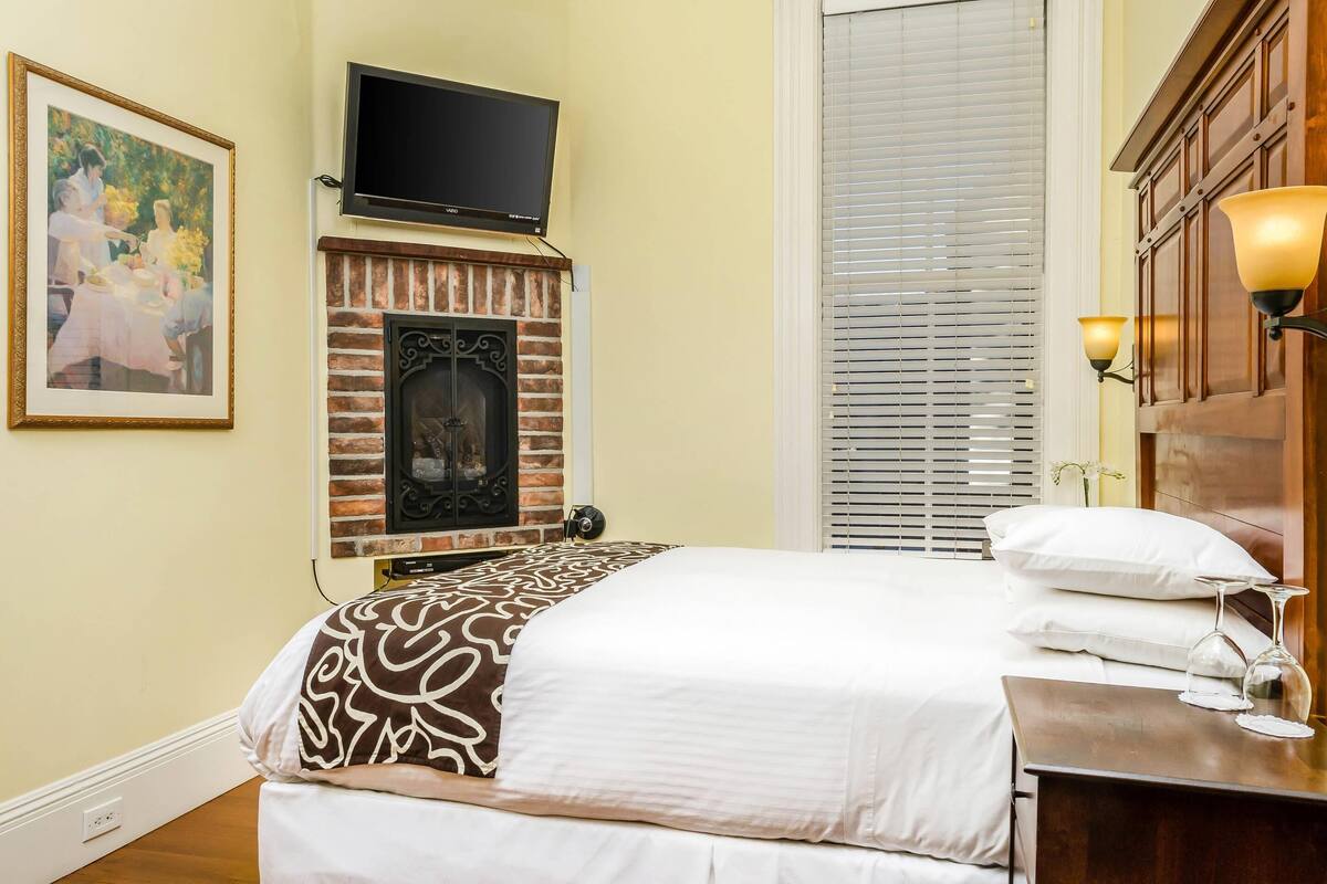 A hotel room with a bed, a fireplace, a television, and a window with blinds.