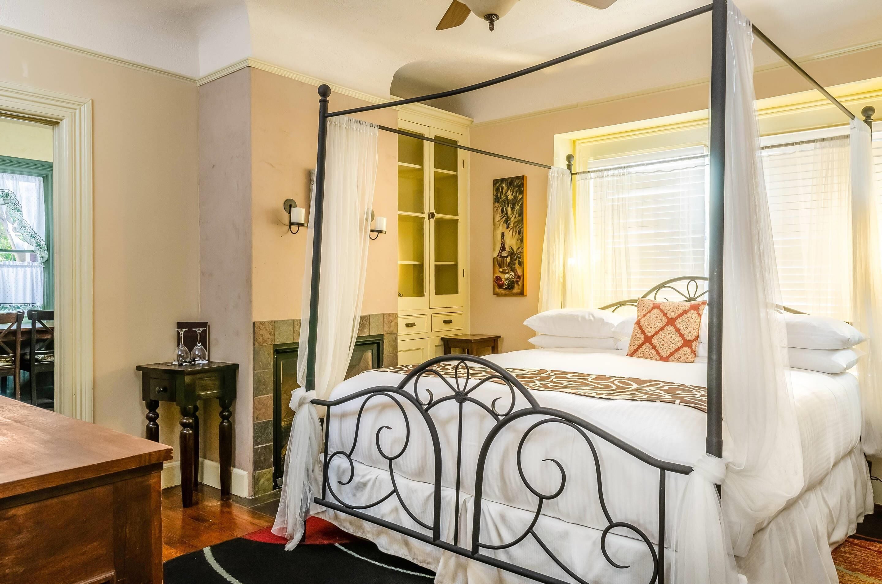 A four-poster bed with a canopy, a wooden nightstand, a small table with a lamp, and a painting on the wall.