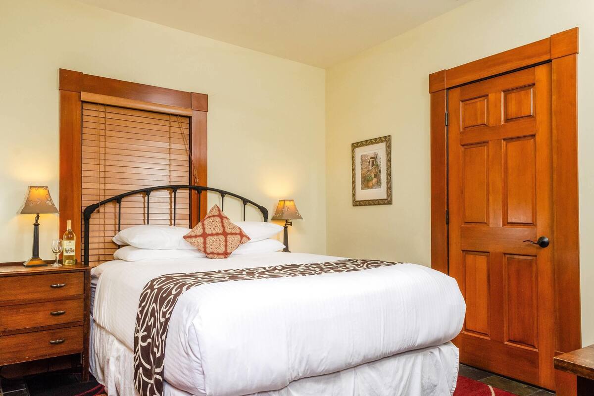 A neatly made bed with a patterned bedspread, a wooden headboard, two bedside lamps, a wooden nightstand, and a wooden door.