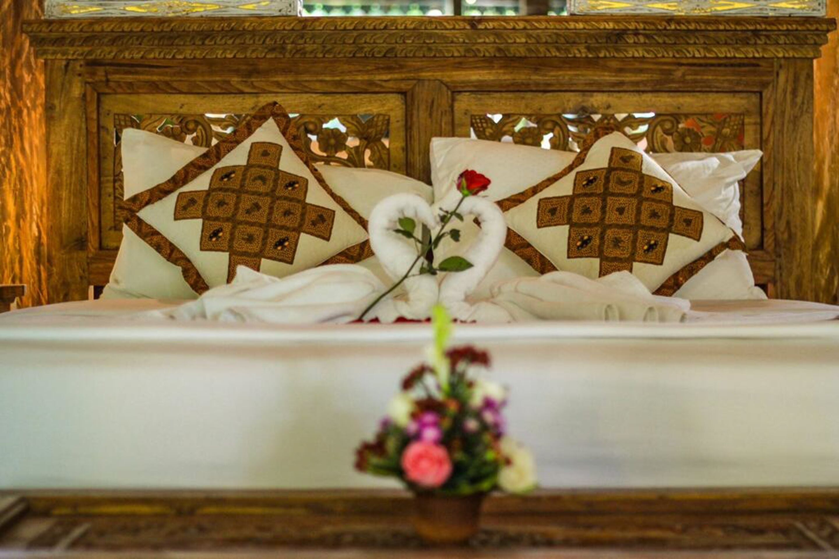 Una cama con cabecera de madera, dos almohadas con estampado geométrico, una rosa y un pequeño ramo de flores.