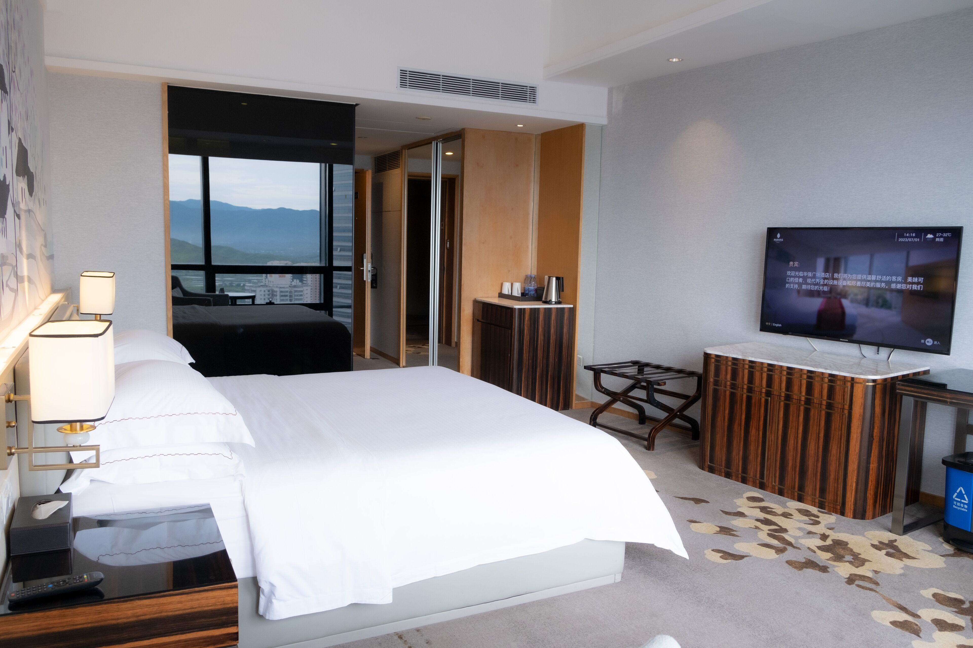 A hotel room with a bed, a desk, a television, and a view of mountains.