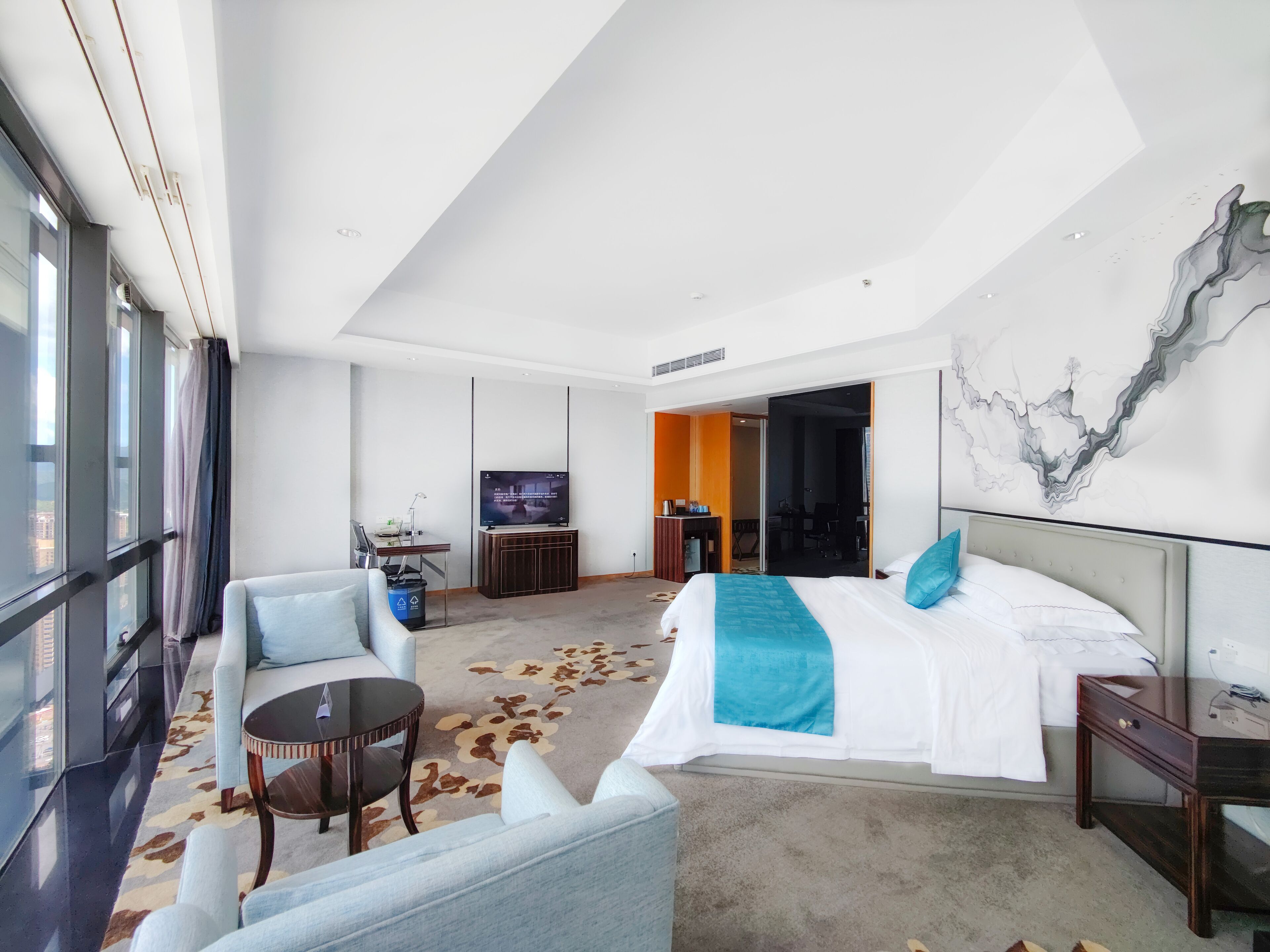 A modern hotel room with a large bed, a desk, a TV, and a view of the city through large windows.