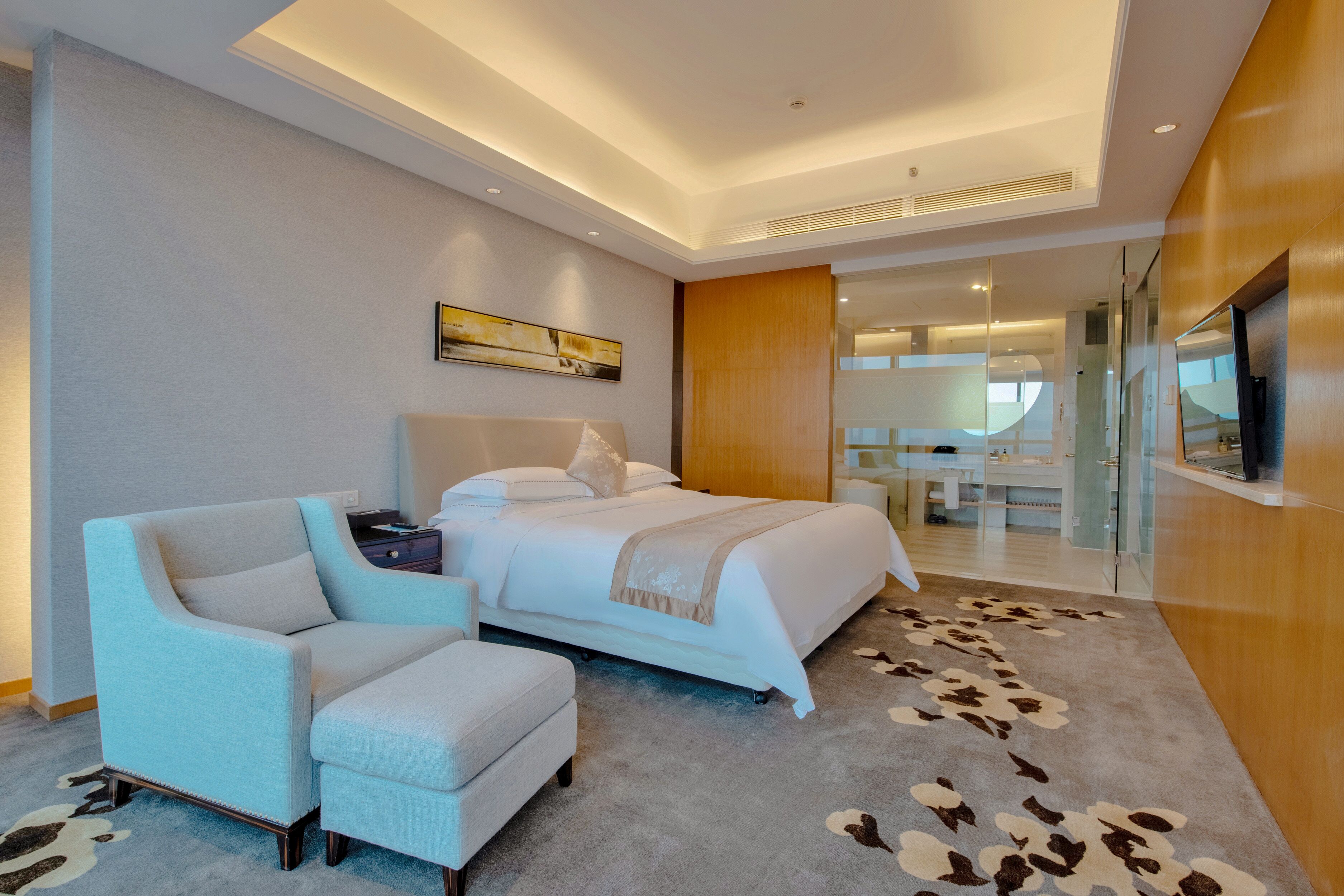 A modern hotel room with a bed, a seating area with a chair and ottoman, a flat-screen TV, and a glass partition separating the room from the dining area.