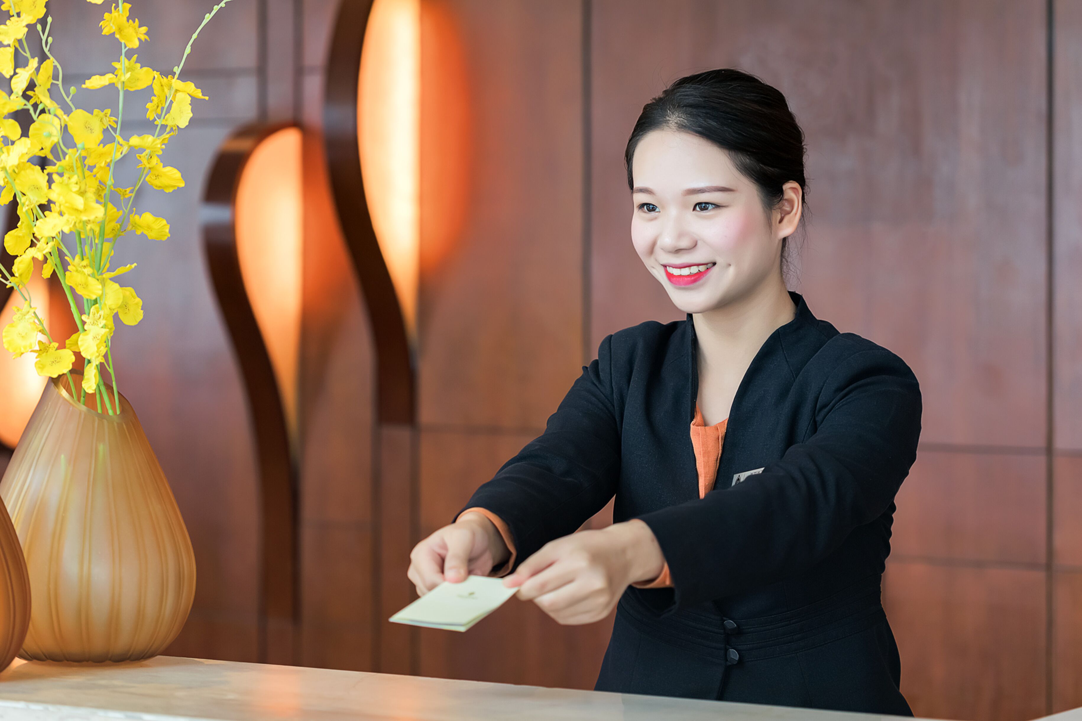 A person handing over a card to another person at a reception desk with a vase of yellow flowers and a wooden wall in the background.