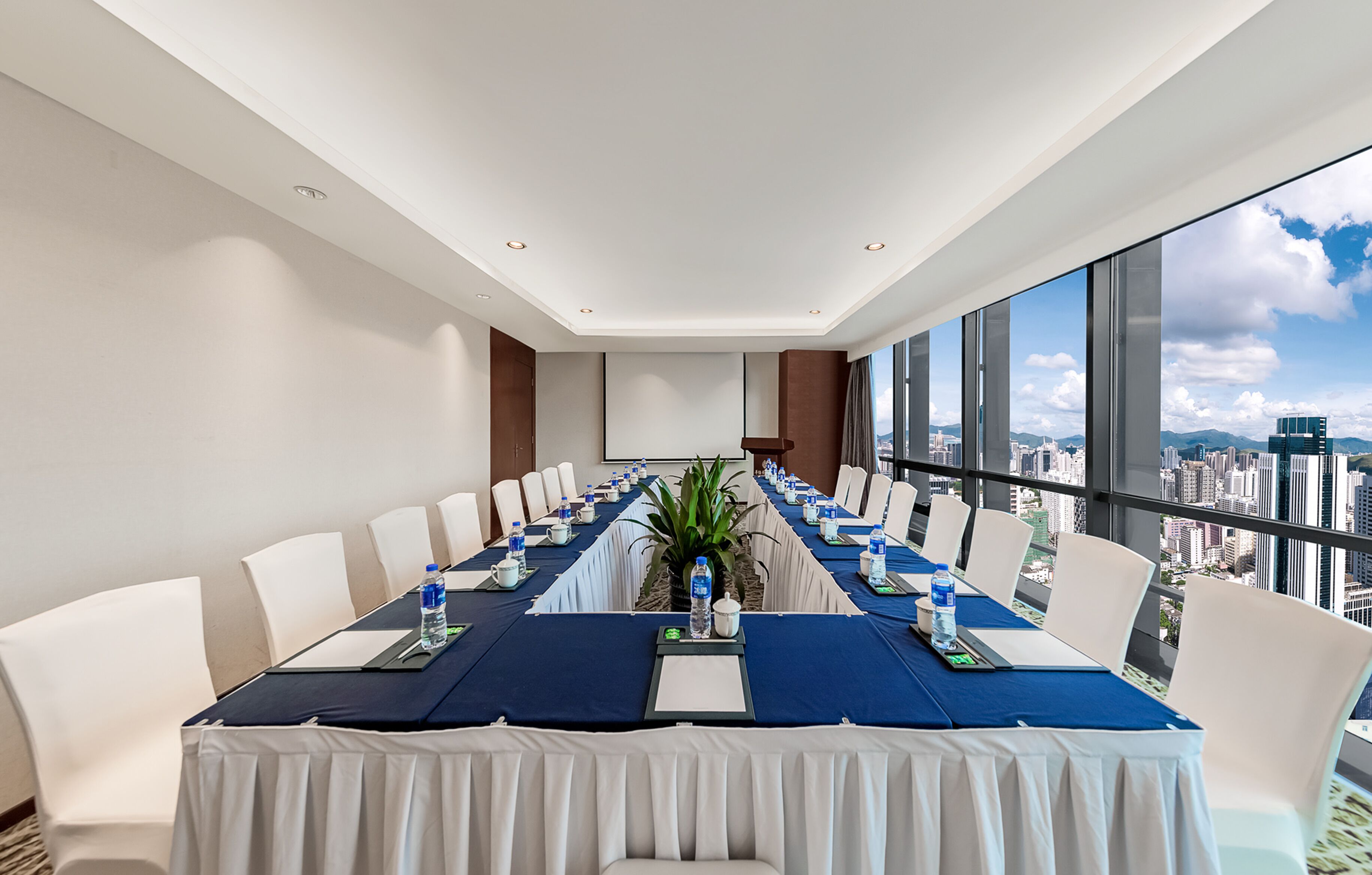 Un salón de conferencias con una larga mesa, sillas blancas y vistas a la ciudad a través de amplias ventanas.
