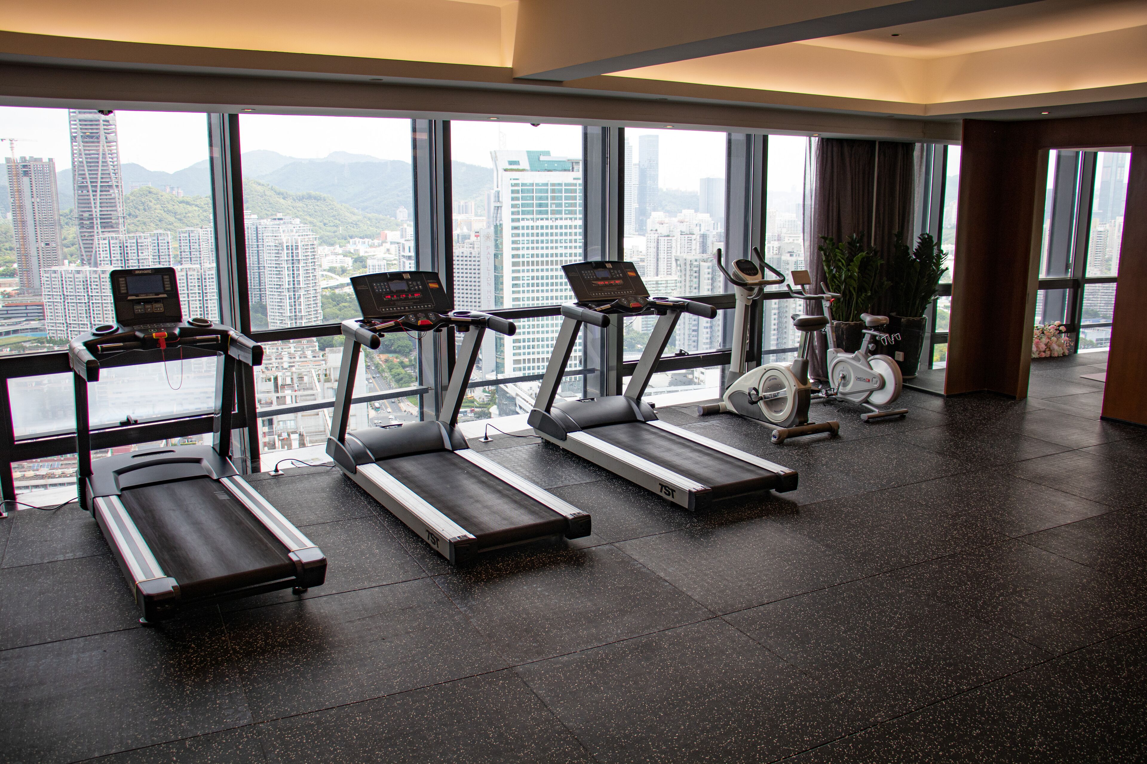 Un gimnasio con cintas de correr, elíptica y bicicleta estática, ventanas amplias con vista a la ciudad.