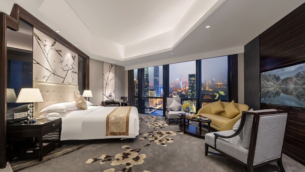 Habitación de hotel con una cama grande, un sofá, una silla, un escritorio y un amplio ventanal con vistas a la ciudad.