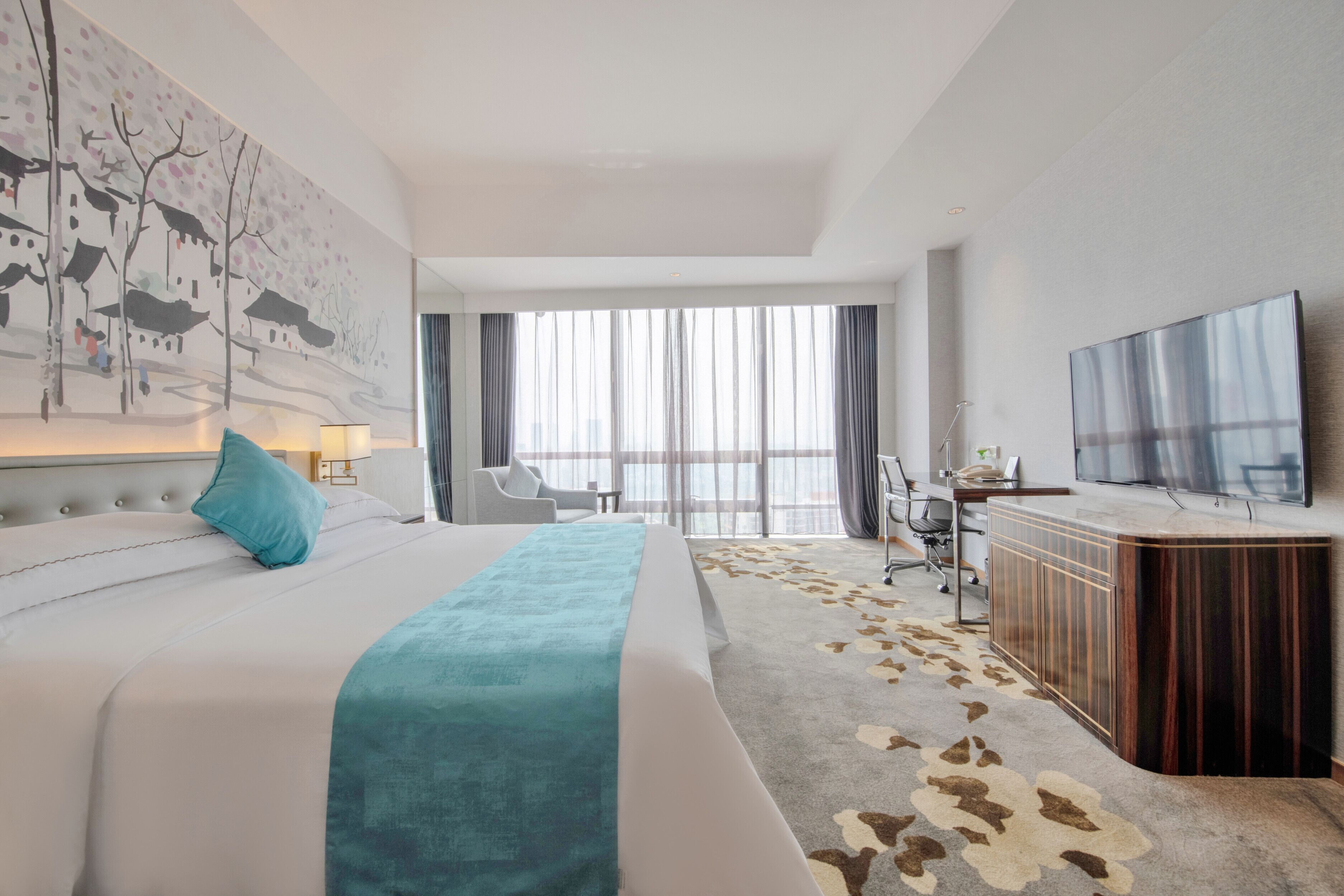 A hotel room with a large bed, a desk with a flat-screen TV, a view of the city through large windows, and a modern decor style.