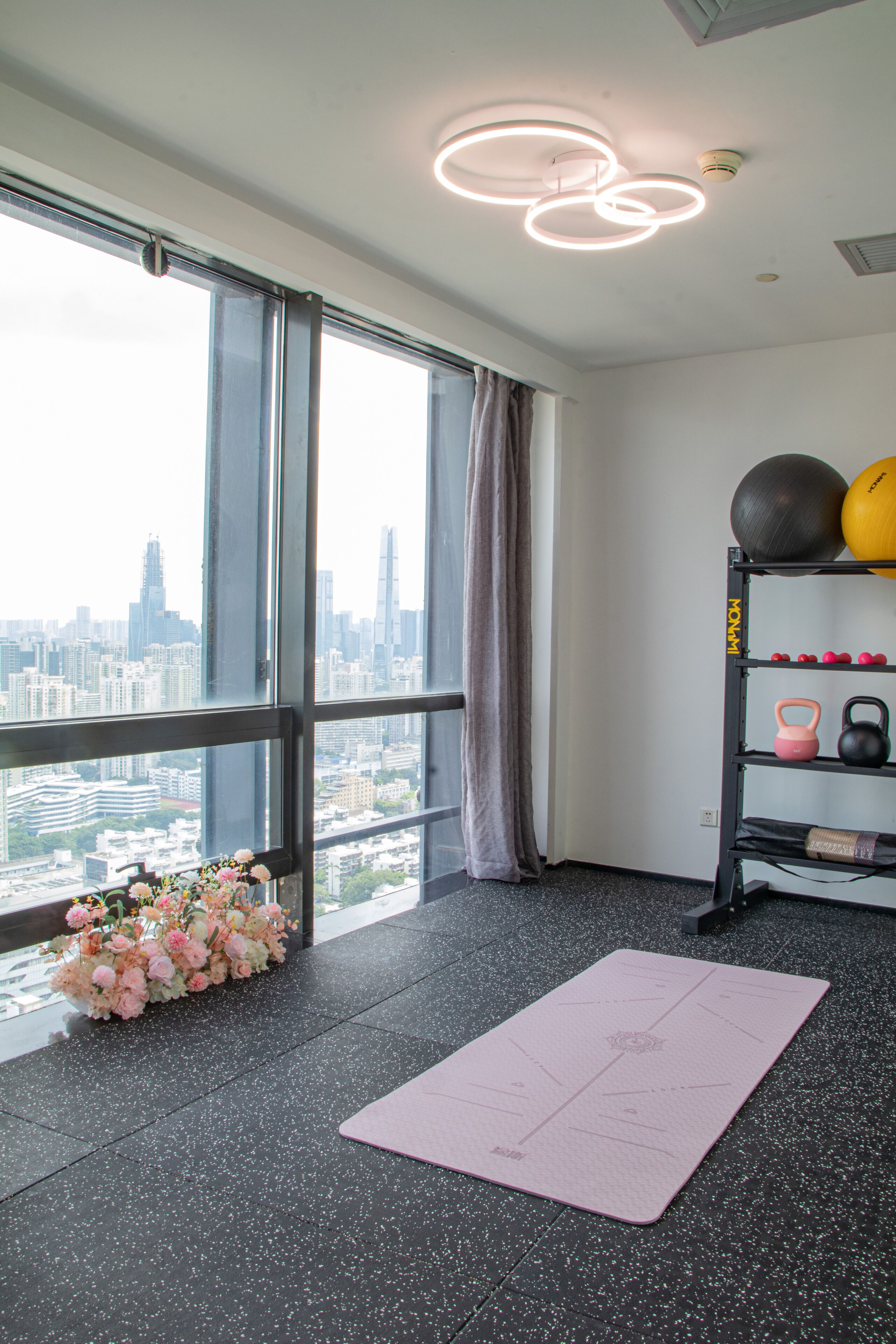 A room with a large window, a pink exercise mat, a shelf with fitness equipment, and a floral arrangement.