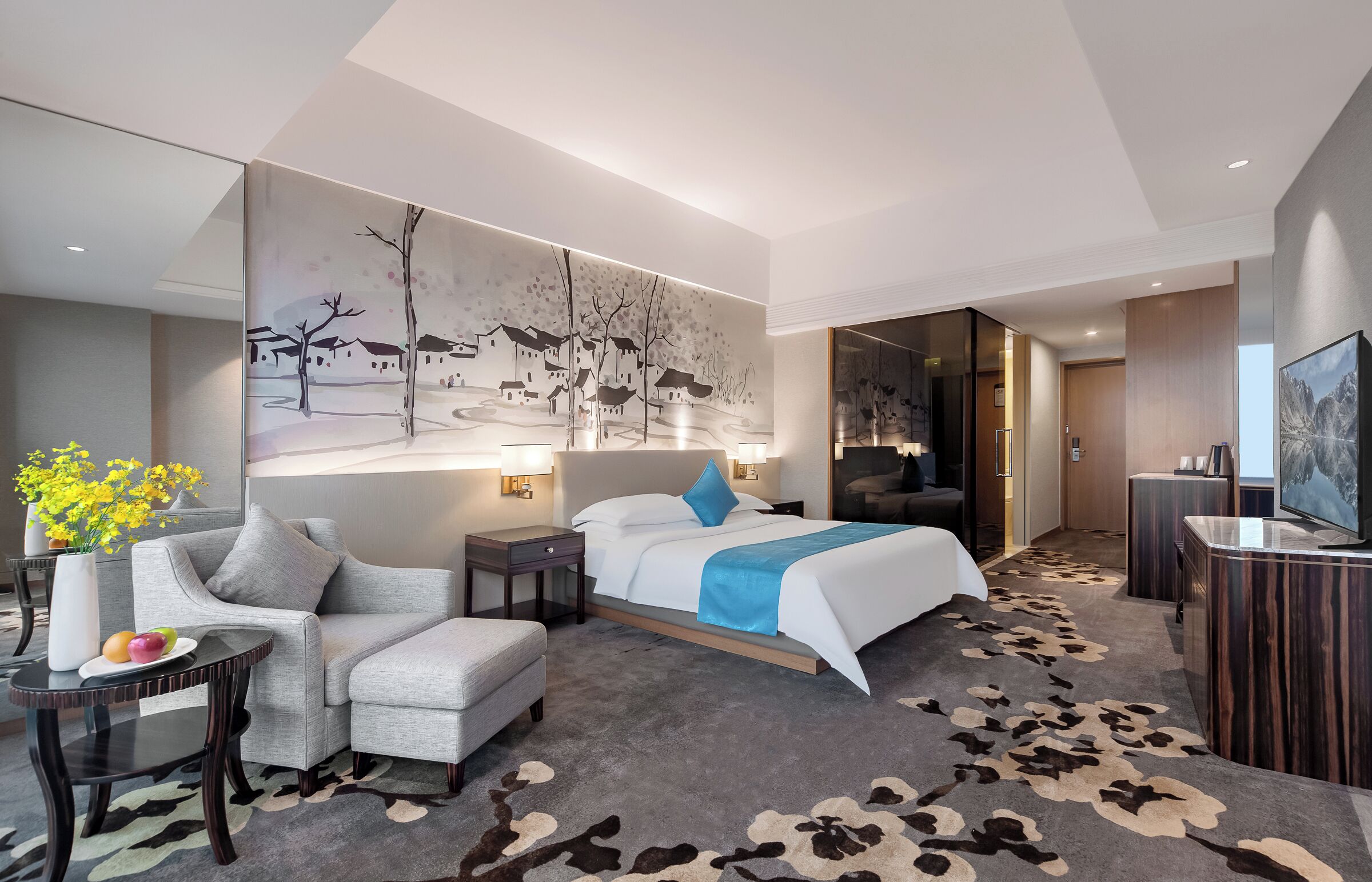 A modern hotel room with a large bed, a sofa, a small table with fruit, a painting on the wall, and a TV in the corner.