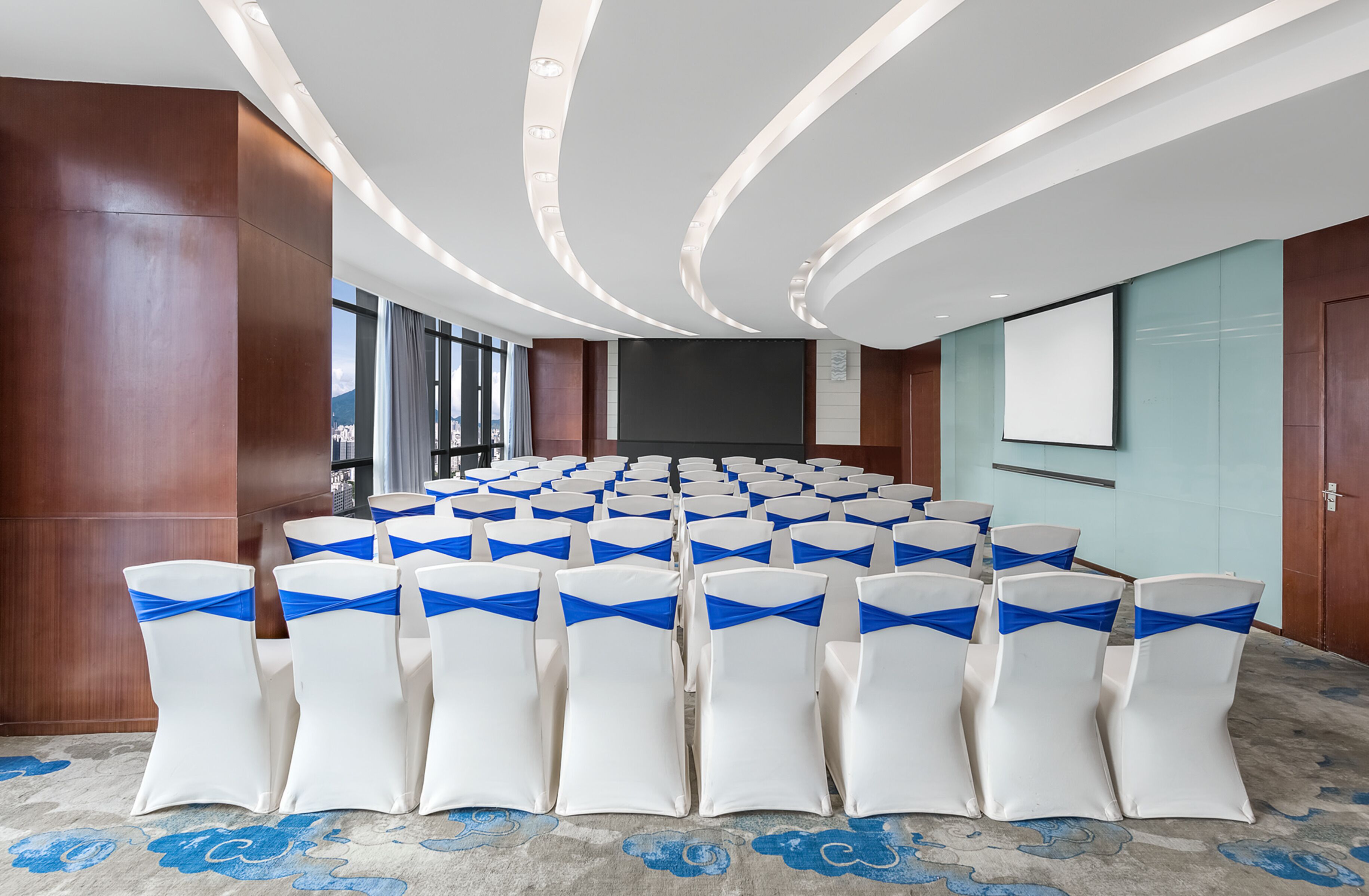 Un salón de conferencias con filas de sillas blancas con cintas azules, una pantalla grande y un diseño de techo moderno.