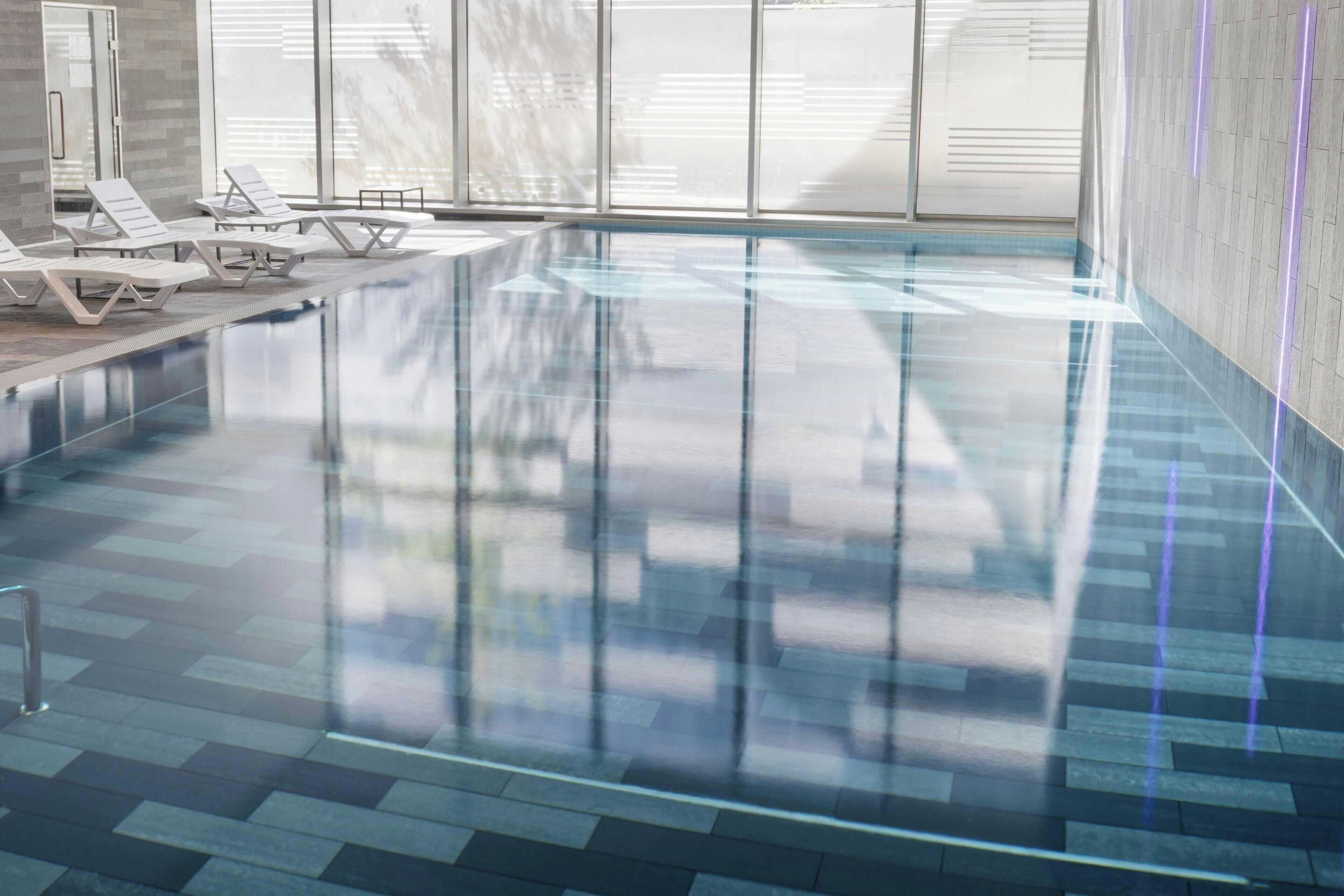 Une piscine intérieure moderne avec des parois en verre transparent, des chaises longues et un sol carrelé.