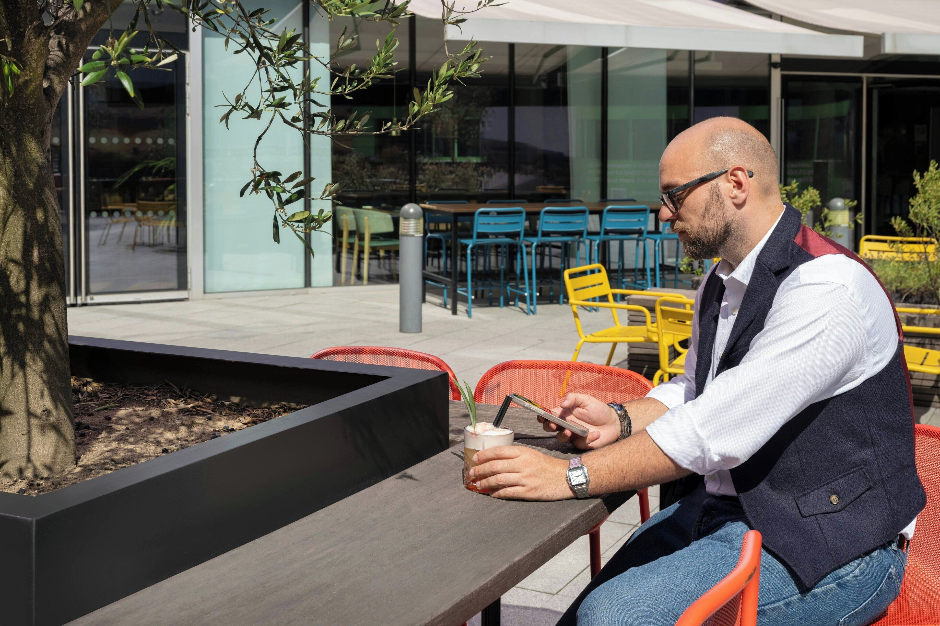 A man sitting at an outdoor table with a drink, surrounded by chairs and a planter.