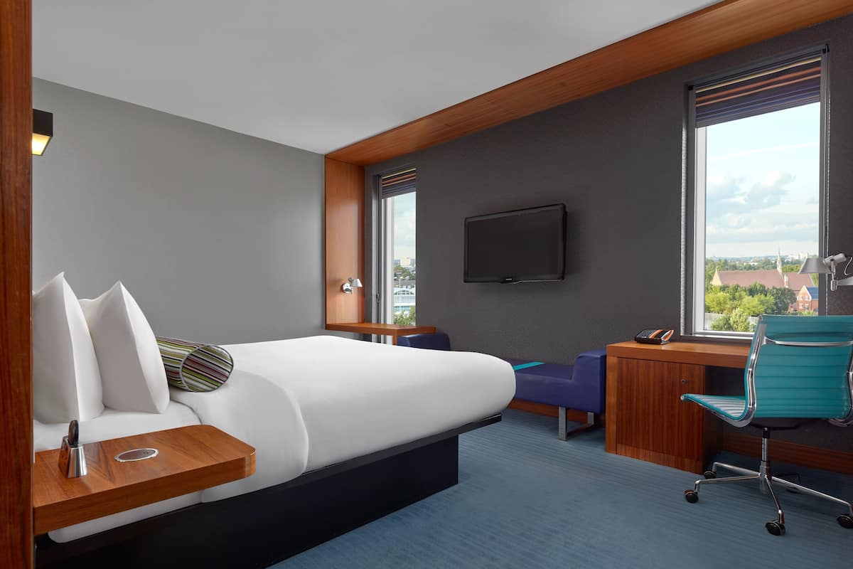 A modern hotel room with a bed, a desk, a chair, a TV, and a window with a city view.