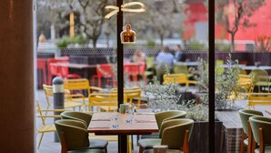 An outdoor dining area with yellow chairs and tables, a view of the street, and hanging pendant lights. - Aloft by Marriott London Excel (London)