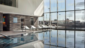 A modern indoor swimming pool with clear glass walls, lounge chairs, and a view of the cityscape.