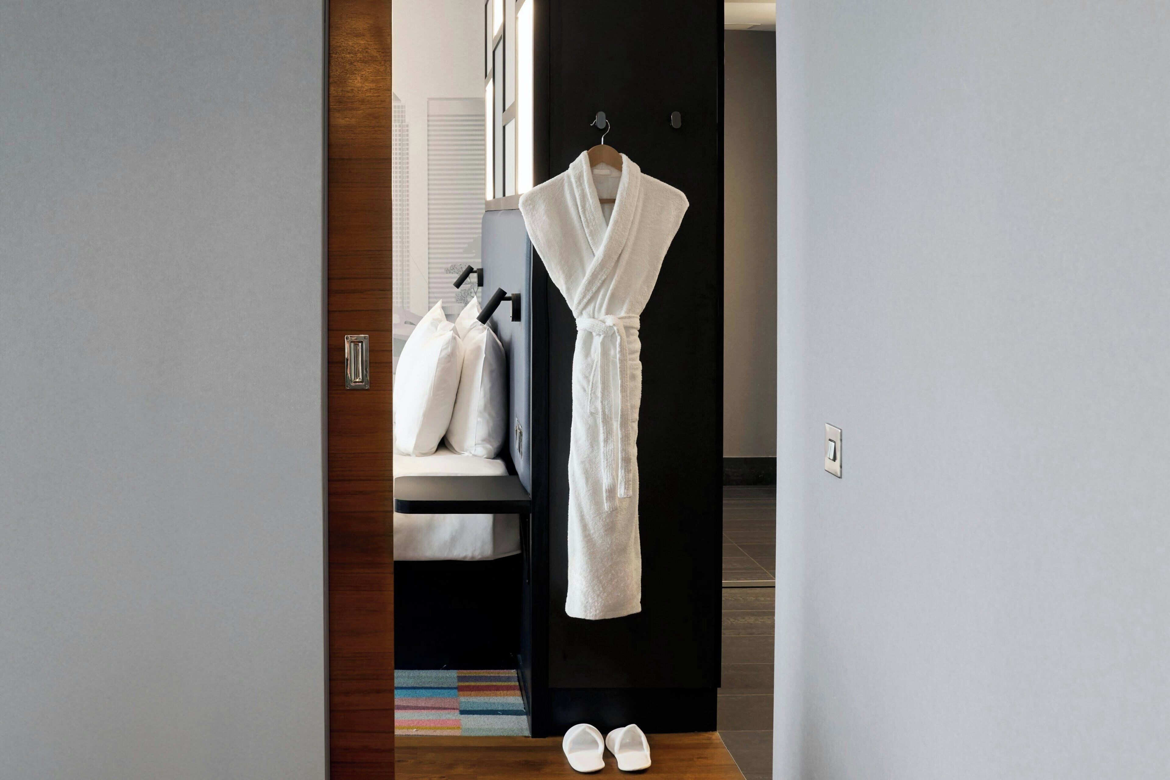 Ein Hotelzimmer mit Holzboden, einem schwarzen Kleiderschrank, einem weißen Bademantel an einem Haken und einem Paar weißer Hausschuhe auf dem Boden.