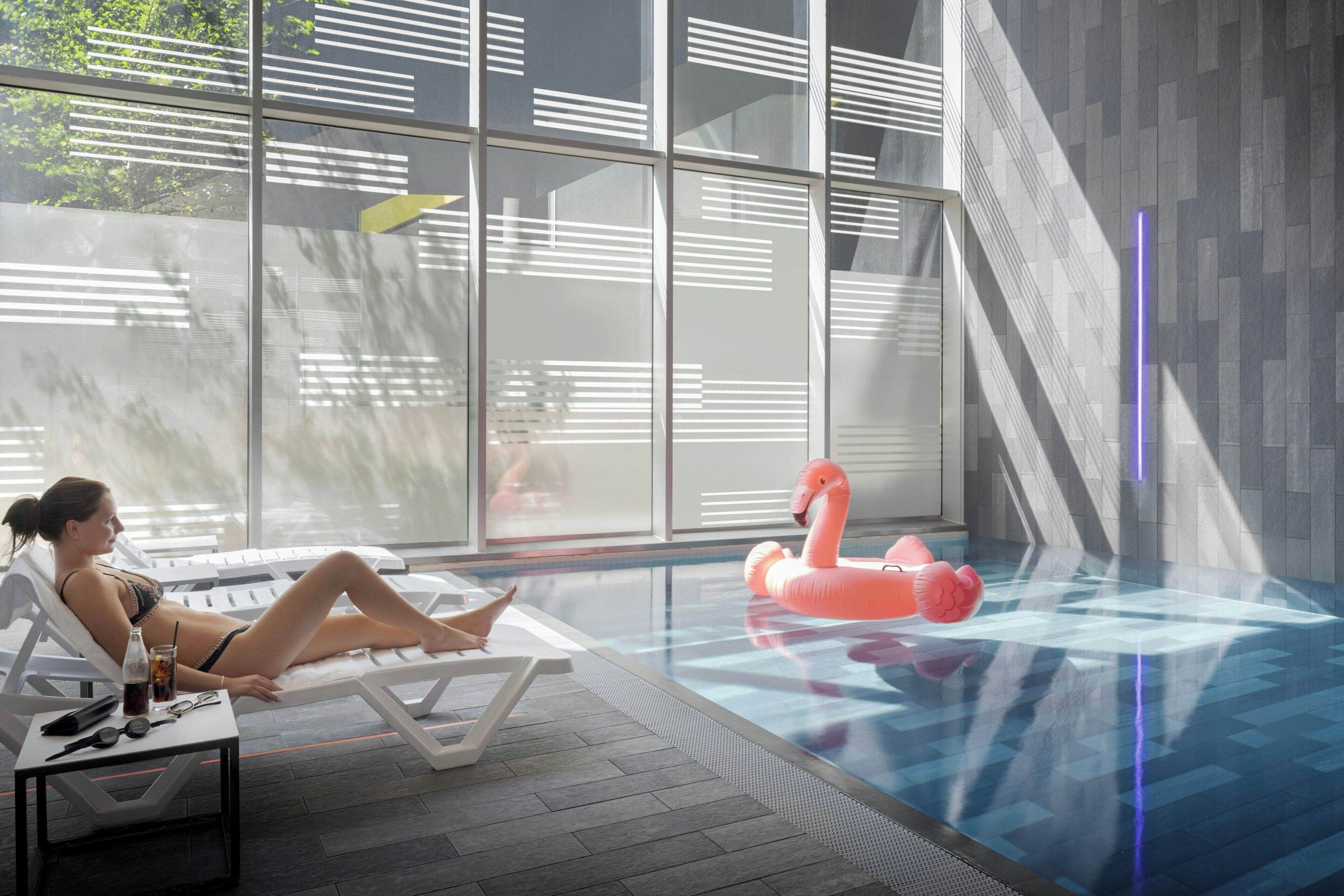 Un área de piscina interior moderna con una barrera de cristal, sillas de descanso y un flotador con forma de flamenco rosa.