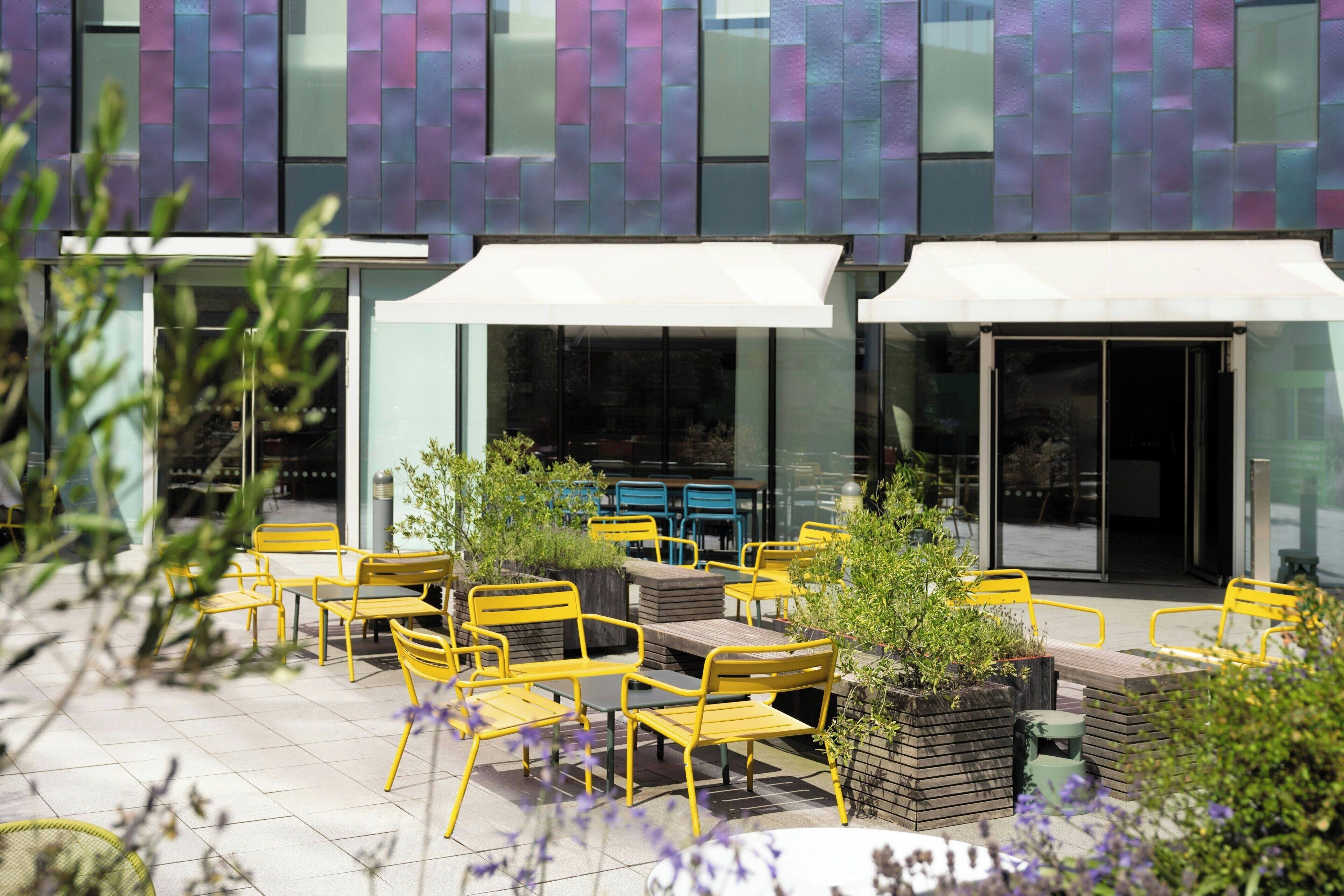 Un espace extérieur aménagé avec des chaises et des tables jaunes, entouré de jardinières et bordé par la façade d’un bâtiment moderne.