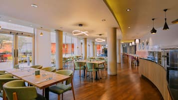 A spacious restaurant with wooden tables and green chairs, large windows, and modern pendant lighting.