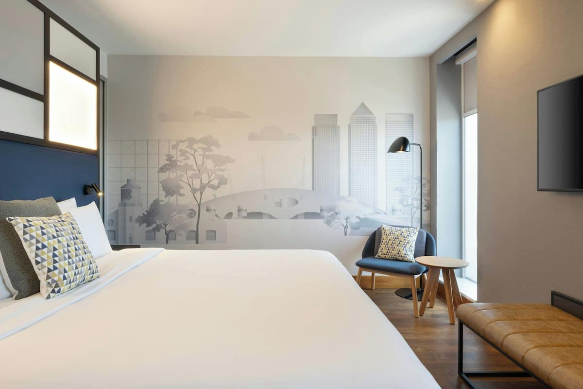 A modern hotel room with a large bed, a chair, a small table, and a TV mounted on the wall.
