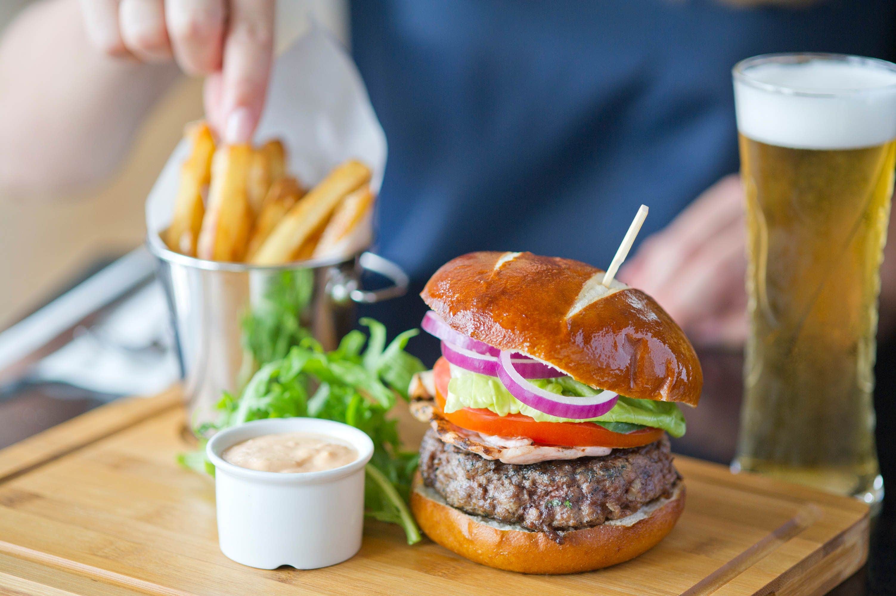Un hamburger garni de laitue, de tomates et d’oignons rouges, un verre de bière et un petit récipient de sauce, le tout présenté sur une planche en bois.