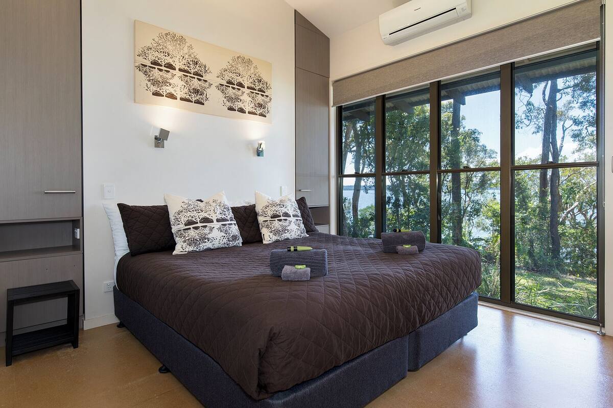Un dormitorio con una cama grande, una mesita de noche, aire acondicionado de pared y un amplio ventanal con vistas a árboles.