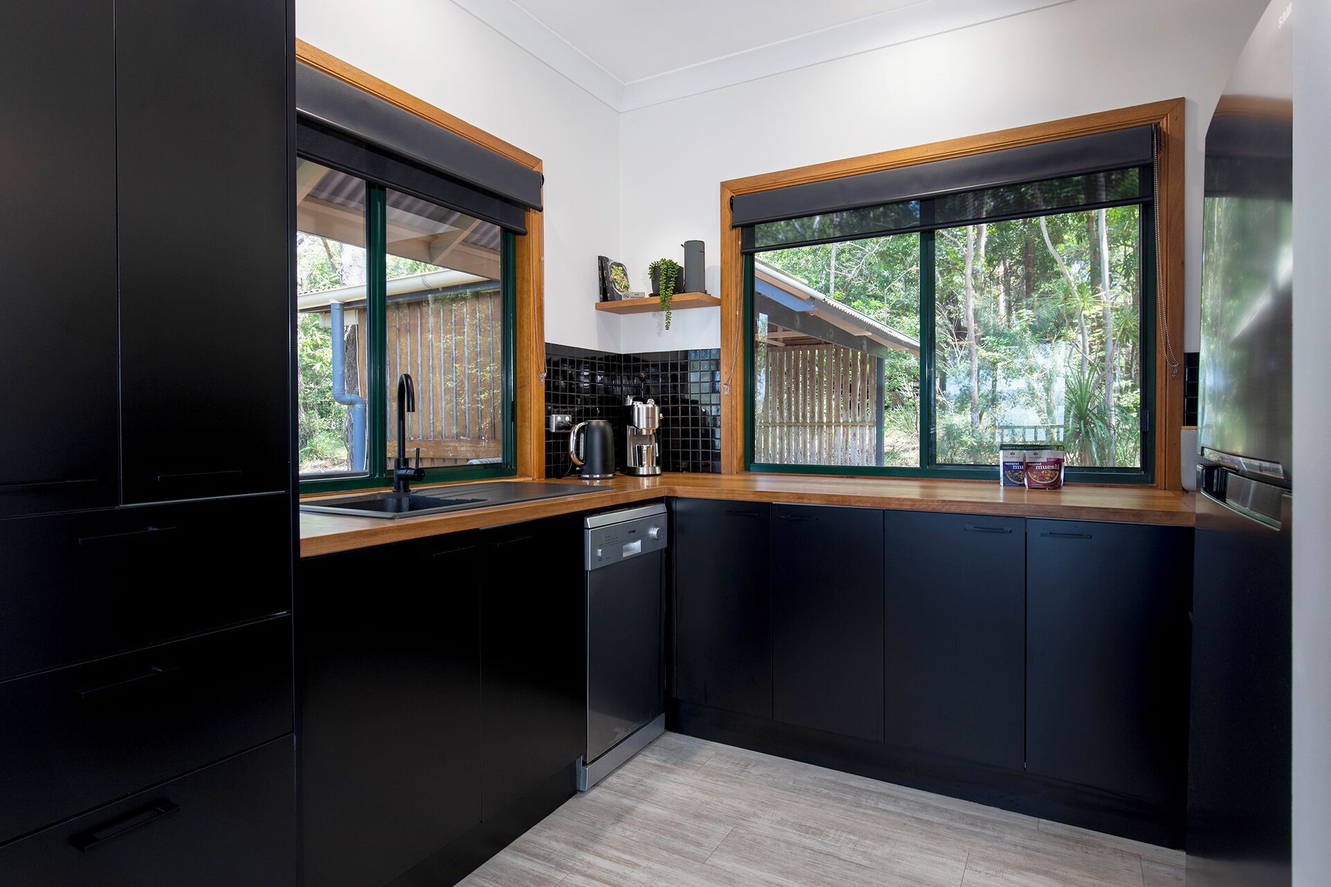 A modern kitchen with black cabinetry, wooden countertops, and large windows offering a view of the outdoors.