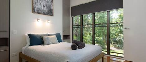 Une chambre avec une grande fenêtre, un lit recouvert d’une couette blanche et un tableau accroché au mur.