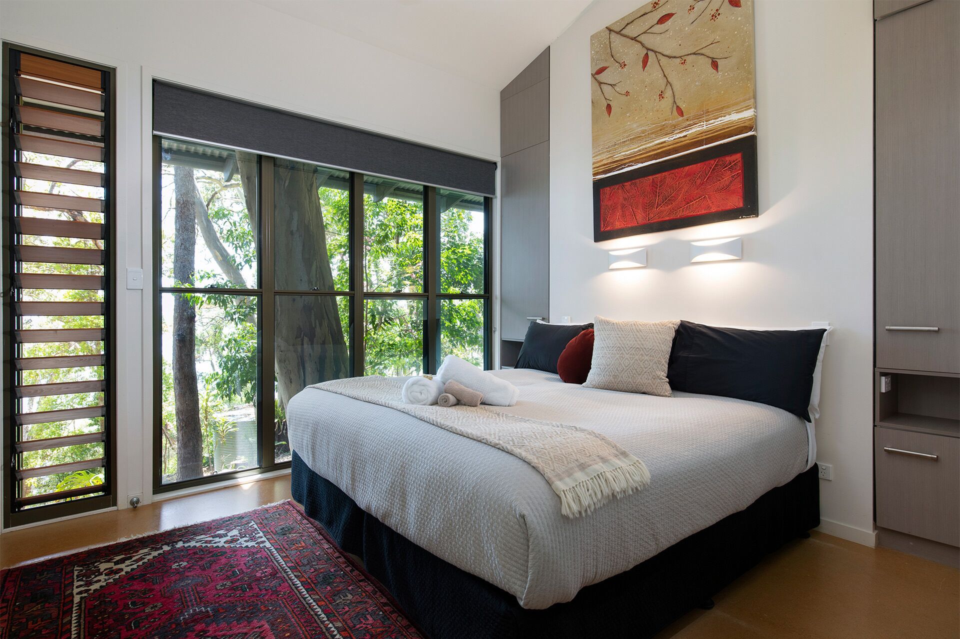 A modern bedroom with a large bed, a view of trees through floor-to-ceiling windows, and a minimalist decor.