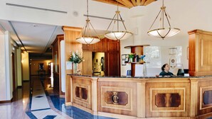 Zona de recepción de hotel con piso de mármol, mostrador de madera e iluminación colgante.