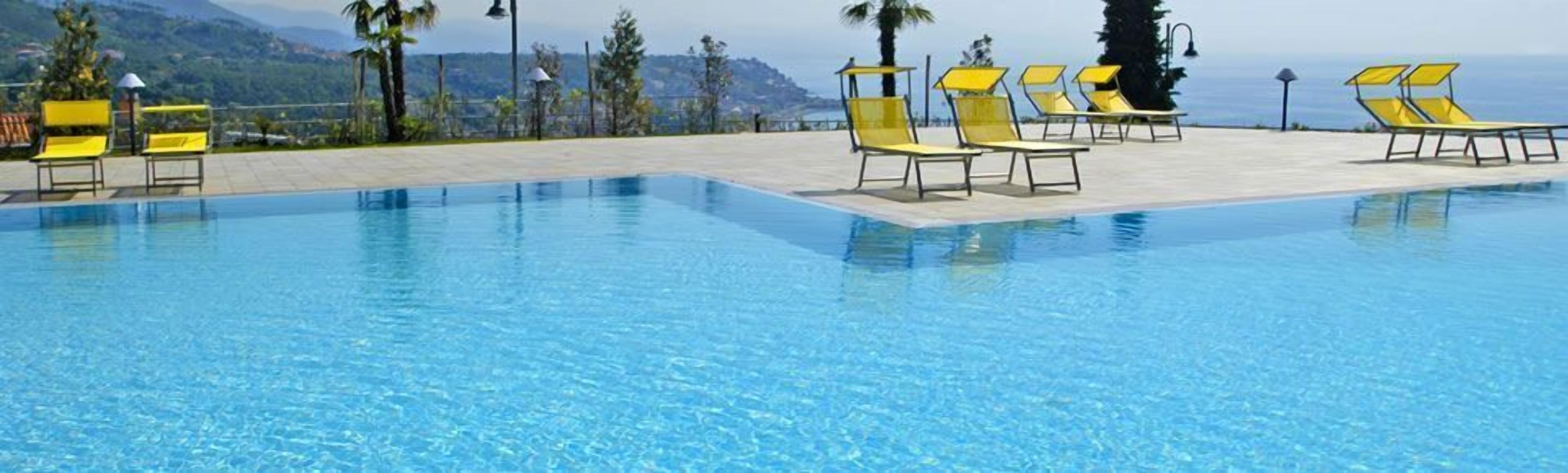 A clear blue swimming pool with yellow lounge chairs and a view of the sea and mountains in the background.