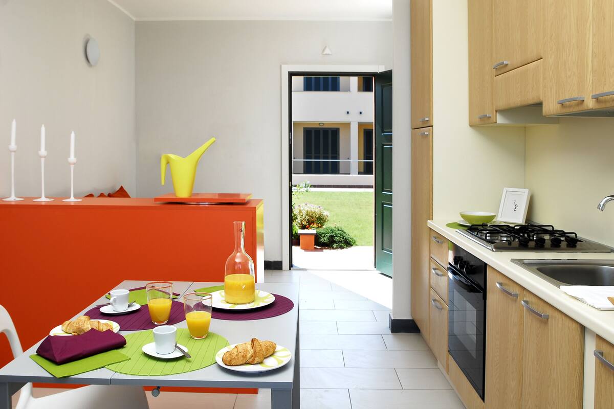 A modern kitchen with a dining area, featuring a table set for breakfast with croissants, juice, and a yellow chair.