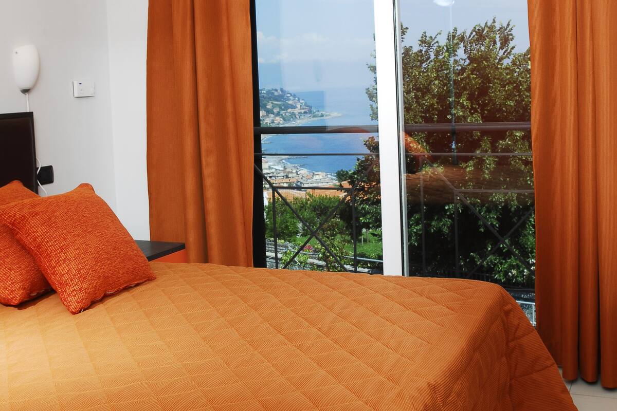 A bedroom with a large bed, orange bedding, and a view of the sea through floor-to-ceiling windows.