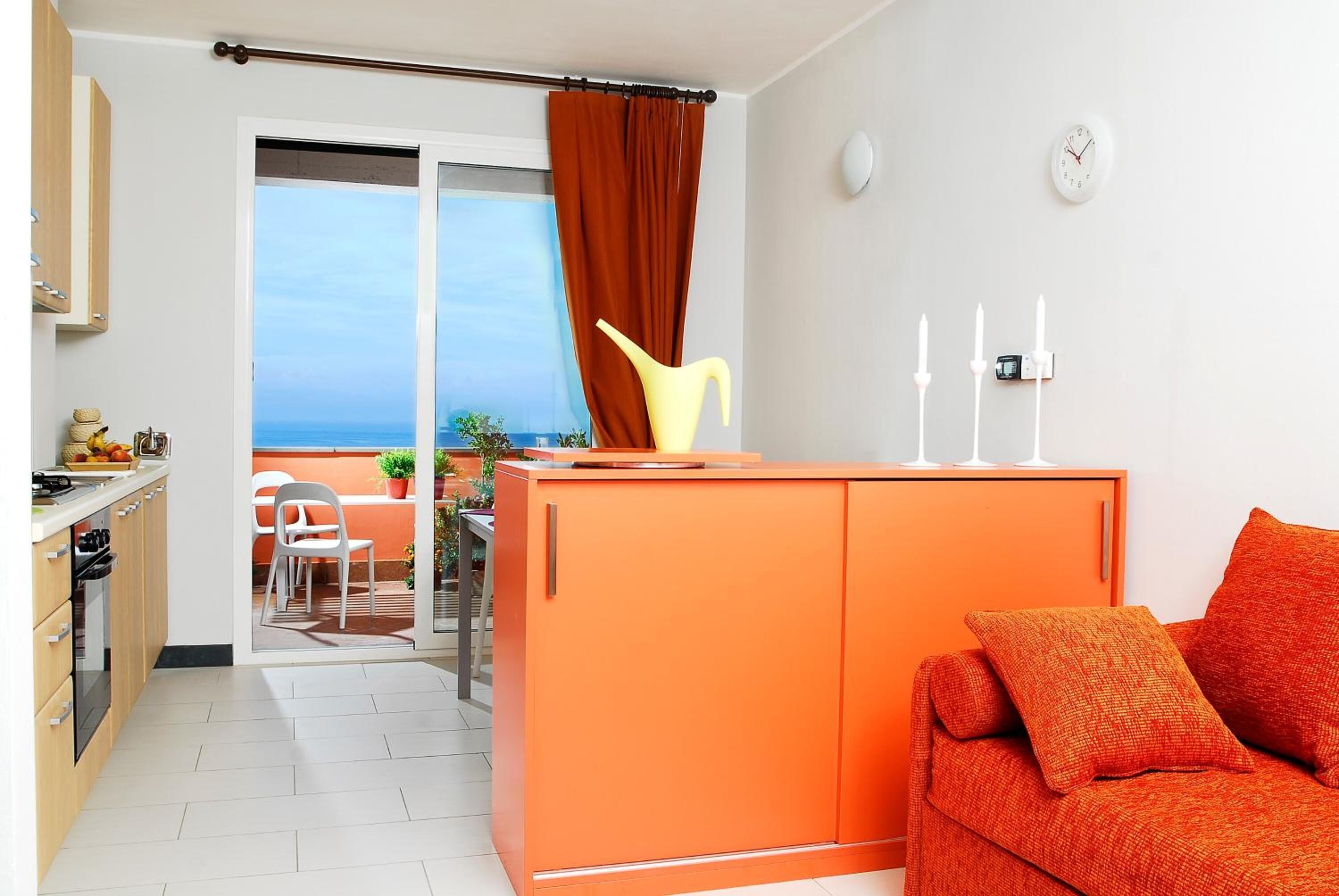 Eine moderne Wohnung mit einem orangefarbenen Sofa, einem Essbereich mit Tisch und Stühlen, einer Küche mit Schränken und Meerblick durch eine Schiebetür.